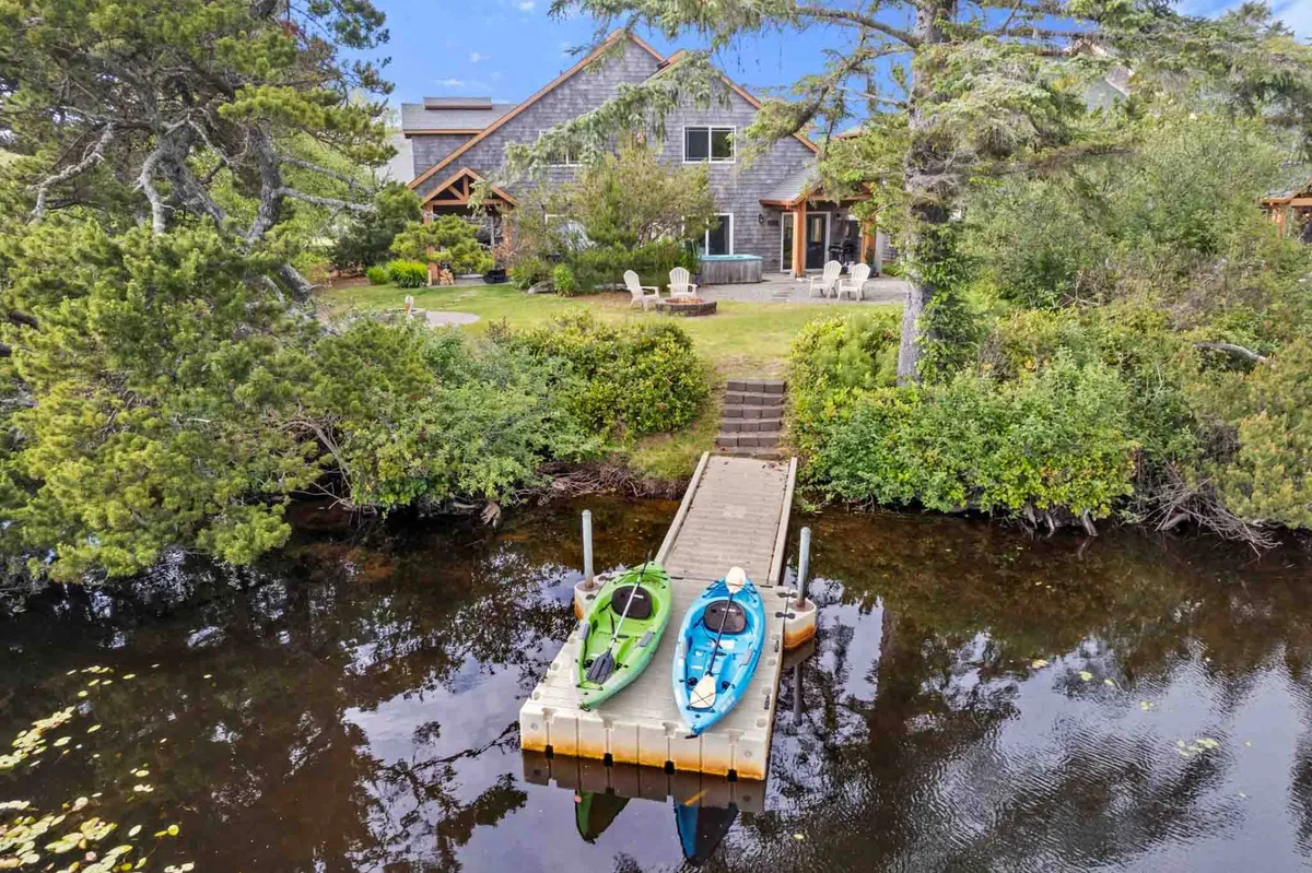 An exterior shot with a dock, kayaks, the house in the background, and surrounding trees and garden. The scene is peaceful, showing not just the dock and kayaks but also the spacious outdoor area leading down to the water—ideal for playing, relaxing, or gathering.