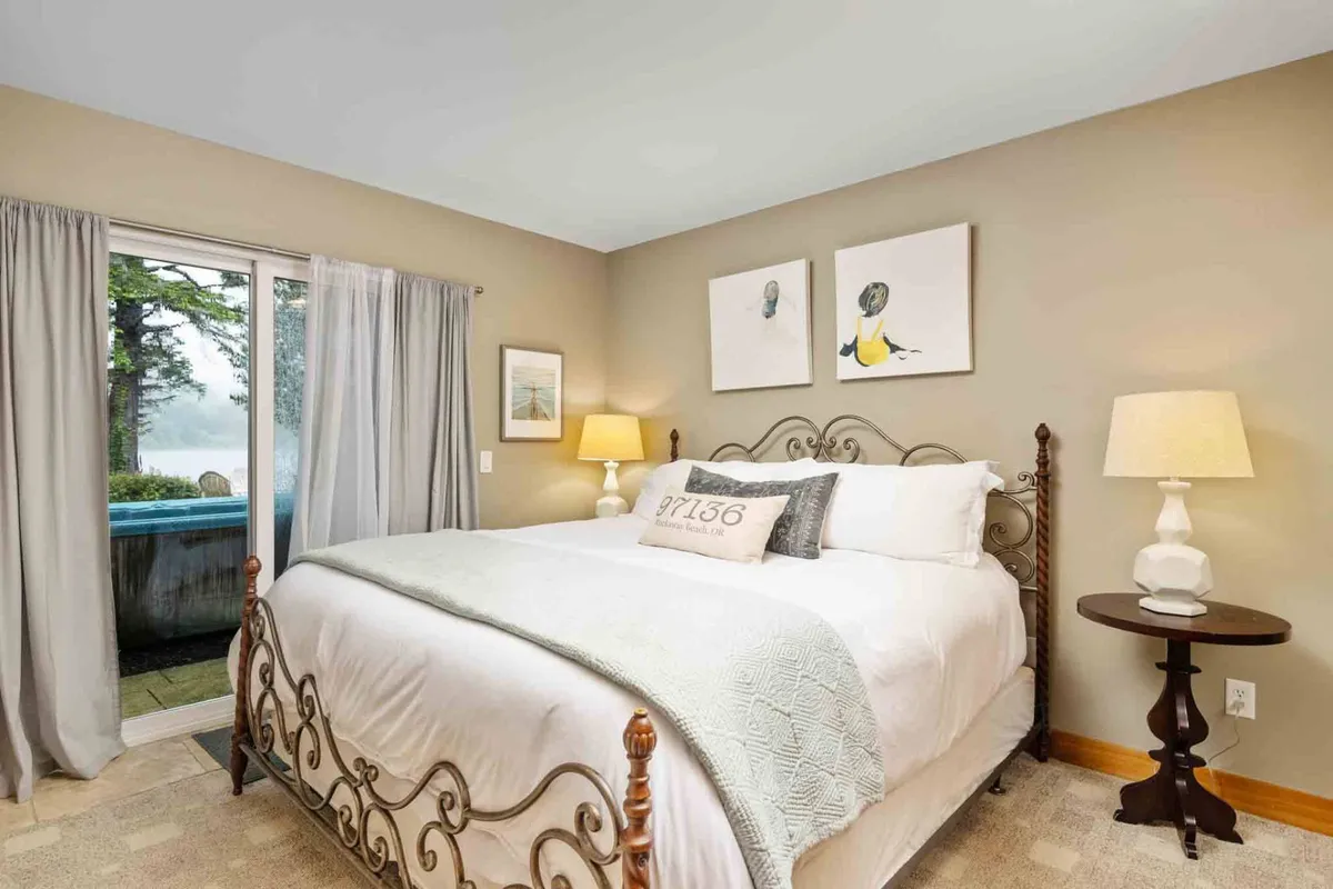 A cozy bedroom featuring a comfortable bed with decorative pillows, framed artwork above the headboard, a bedside lamp, and curtains near a window. The overall ambiance is serene and restful, perfect for recharging.