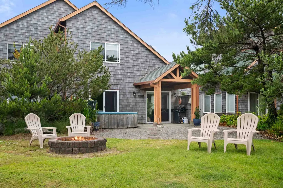 This exterior image showcases a cozy fire pit surrounded by lawn chairs, with a hot tub visible nearby and the home's shingle exterior and landscaping in the background. The setup highlights spaces for both relaxation and social gatherings, all within a natural setting.