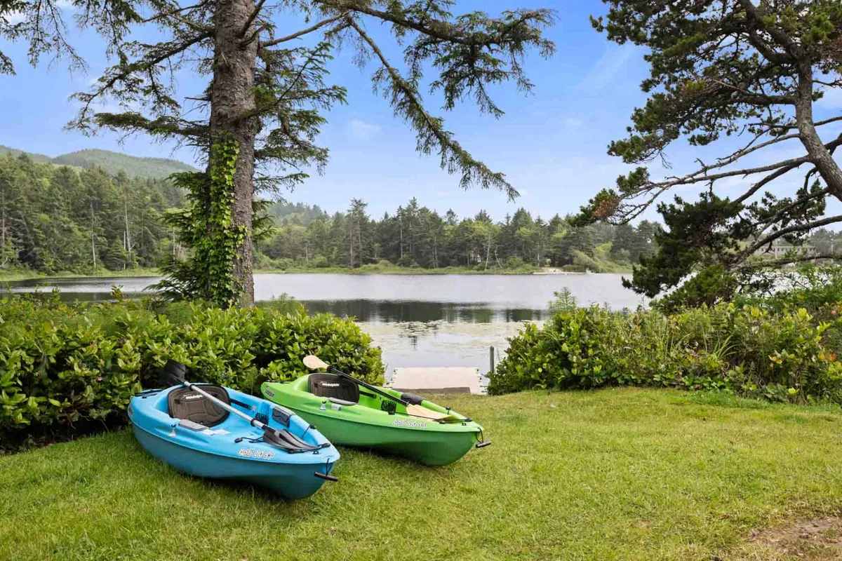 A serene view of two kayaks, one blue and one green, resting on the shore of a calm lake, surrounded by lush trees and greenery. The water reflects the peaceful ambiance, suggesting early morning or sunset light. The scene invites thoughts of kayaking, nature walks, and quiet wildlife observation.