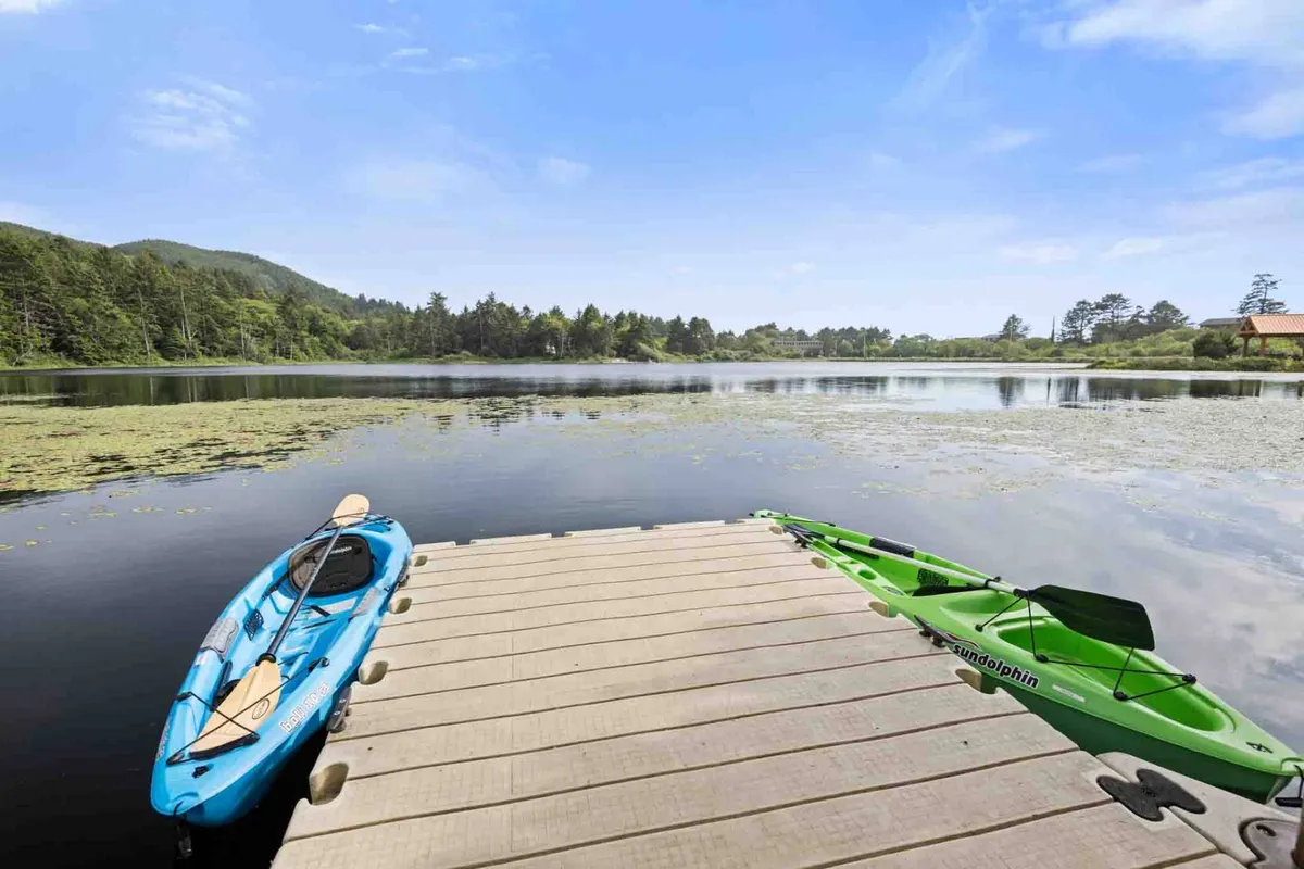 A tranquil lakeside scene featuring two colorful kayaks—one blue, one green—resting on a private wooden dock that extends into the calm waters of Spring Lake. The dock is surrounded by lush greenery and tall trees, evoking a peaceful connection to nature and easy access to lake activities.