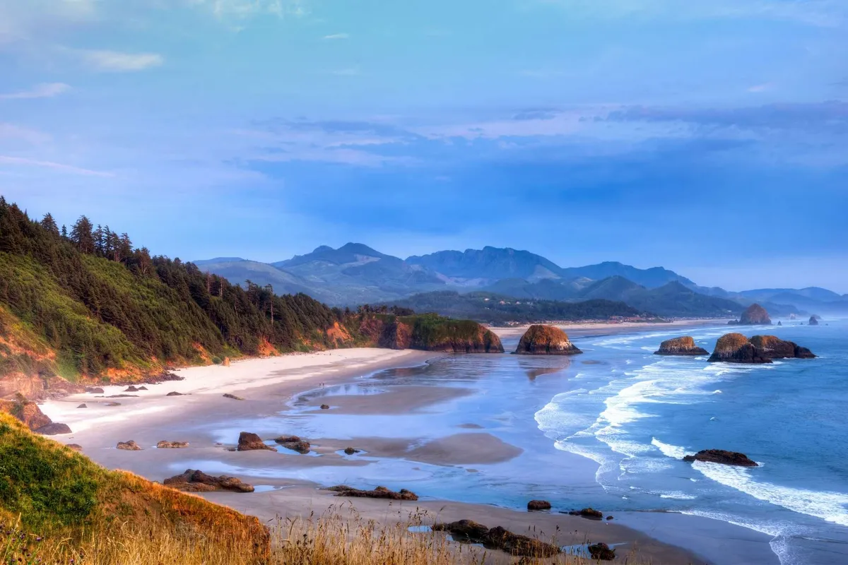 A breathtaking coastal view with rolling ocean waves, dramatic rock formations, distant mountains, and a sandy shoreline. The scene is tranquil and wild, evoking the sense of adventure and awe found on local hikes like those at Neah-Kah-Nie Mountain.