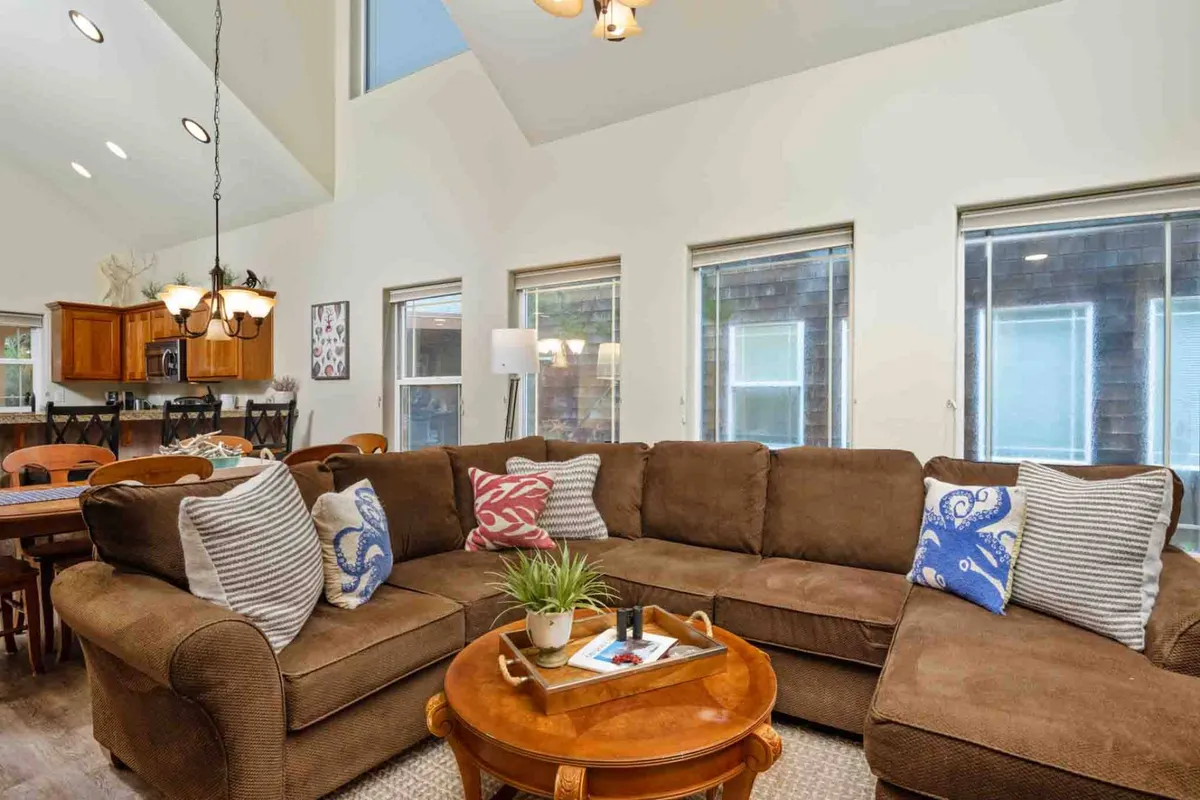 This image captures the heart of the open-concept living space, featuring a large sectional sofa, a coffee table, a view into the dining area, kitchen cabinetry, and expansive windows letting in natural light. The layout is inviting and showcases areas where guests can relax, gather, and enjoy each other's company.