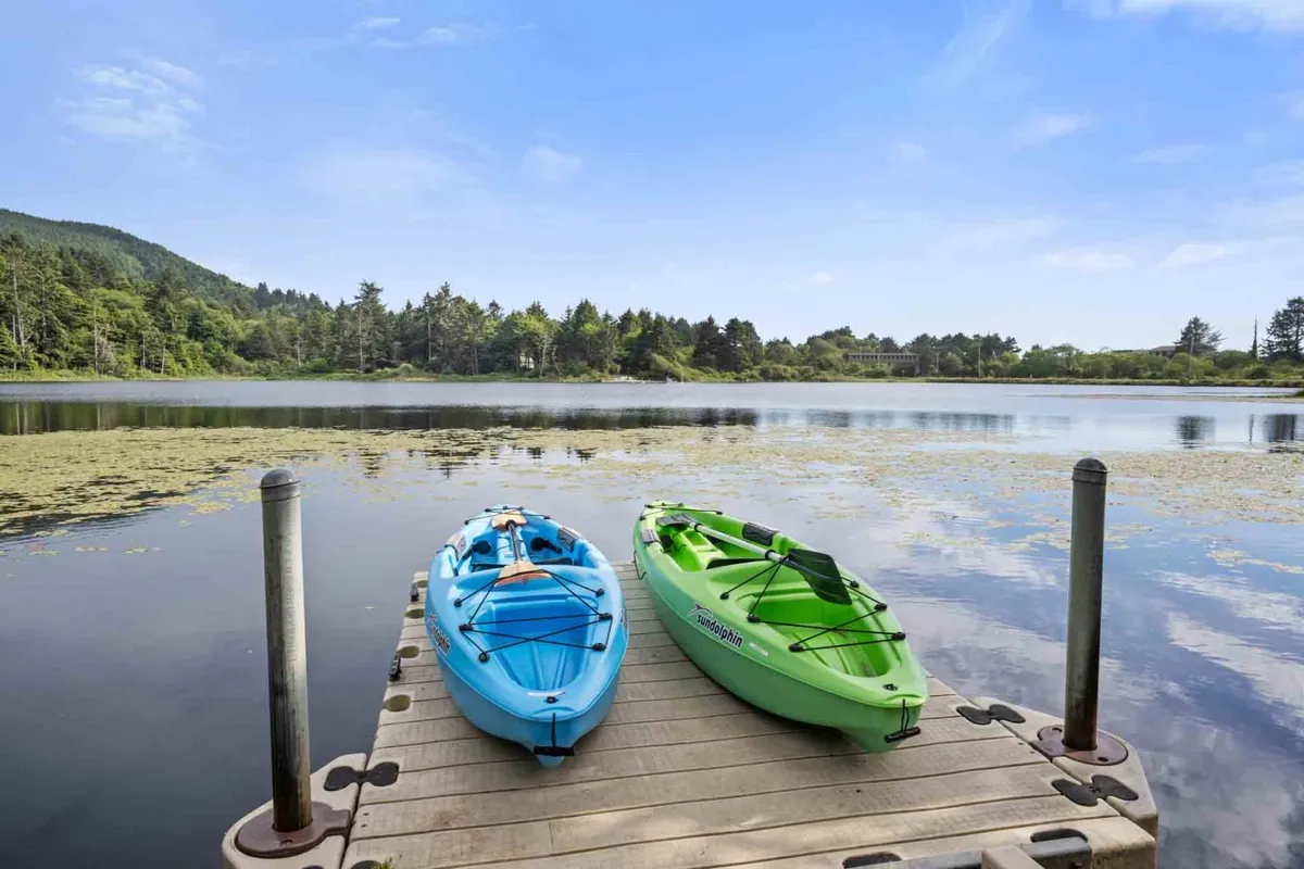 This image shows a serene lakeside scene with a blue and green kayak pulled up next to a private dock, surrounded by calm water and lush trees. The scene is tranquil and inviting with the focus on the dock, kayaks, and the surrounding greenery.