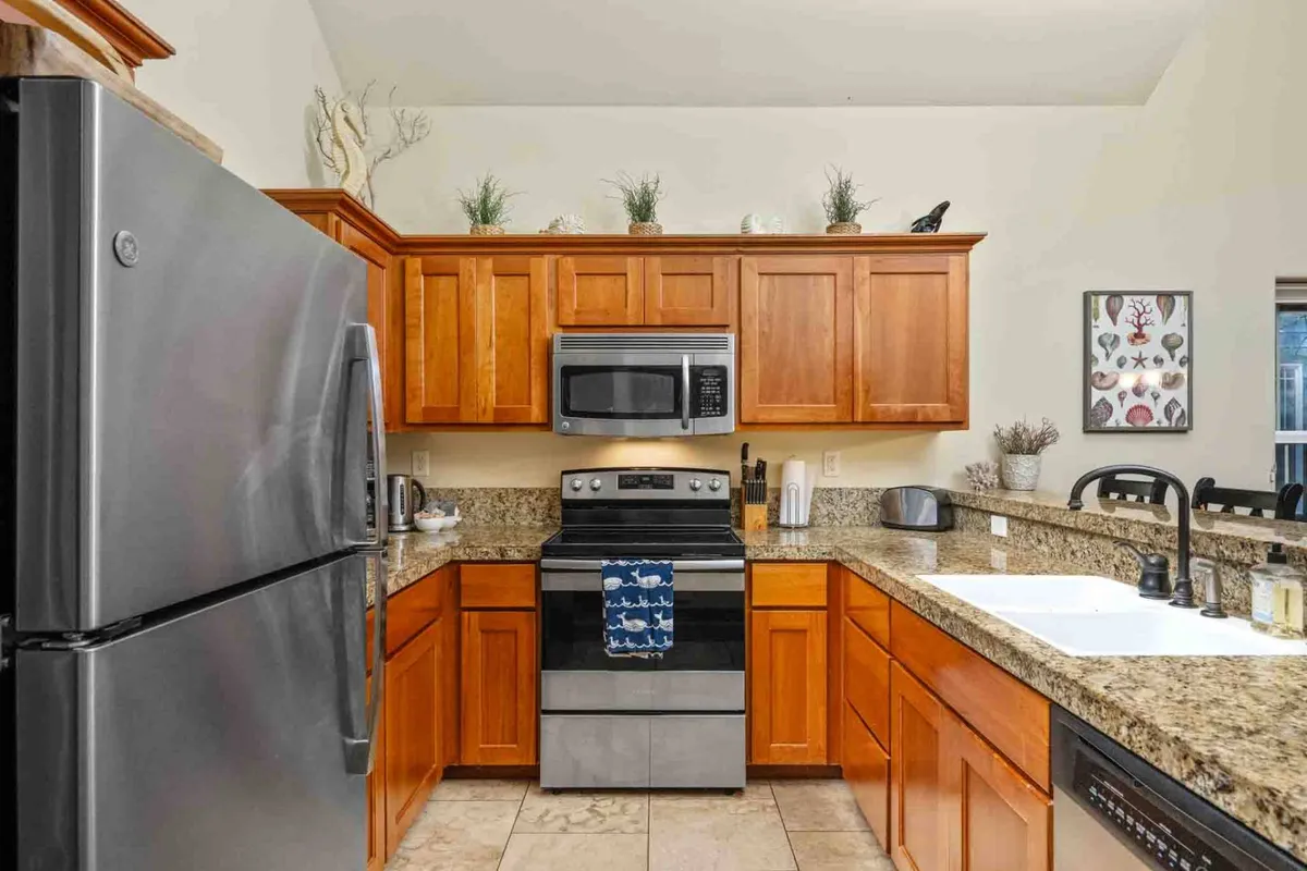 A modern kitchen with stainless steel appliances, granite countertops, wood cabinetry, and decorative wall art, filled with warm, natural light. The image highlights the space where meals are prepared and guests gather for casual chats or to enjoy morning coffee.