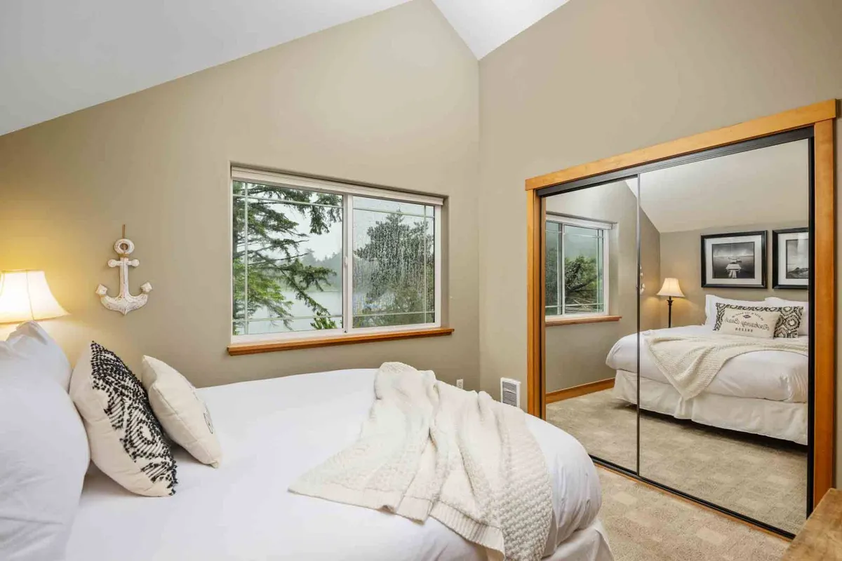 A tranquil bedroom with a bed dressed in white bedding and decorative pillows, a window that frames a view of trees and a tranquil lake, and a mirror reflecting the airy space. Soft light fills the room, emphasizing relaxation and natural beauty.