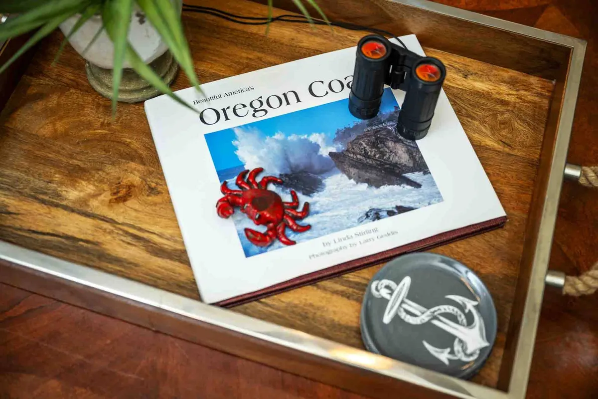A close-up vignette shows a table with an 'Oregon Coast' book, a red crab figurine, binoculars, and a nautical-themed coaster, evoking curiosity about regional attractions and wildlife. The details suggest both planning and reminiscing about local adventures.