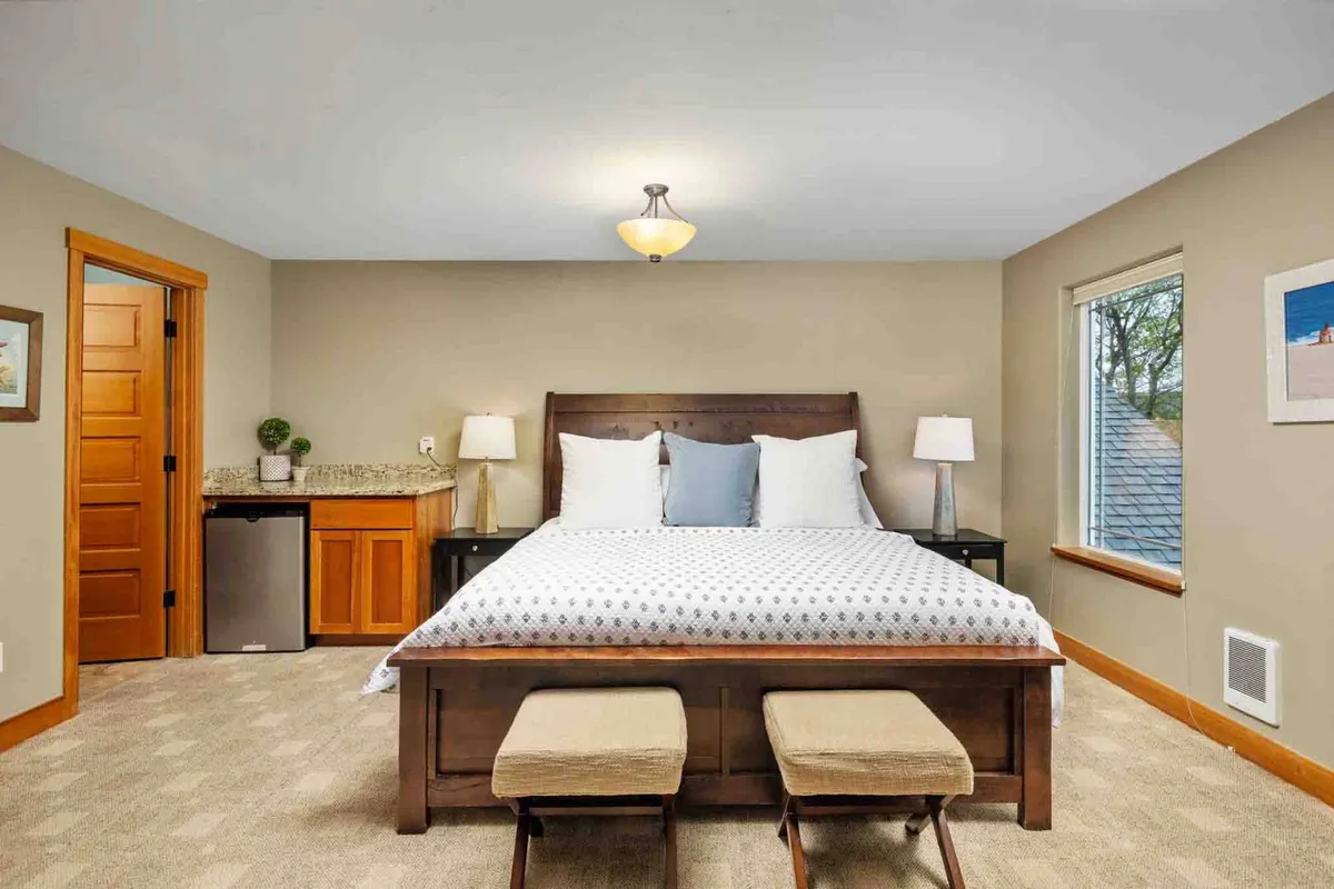 A bright, inviting bedroom featuring a comfortable bed, nightstands, a large window allowing natural light, and a convenient mini-fridge. The mood is cozy and private, ideal for unwinding with a book or morning coffee.